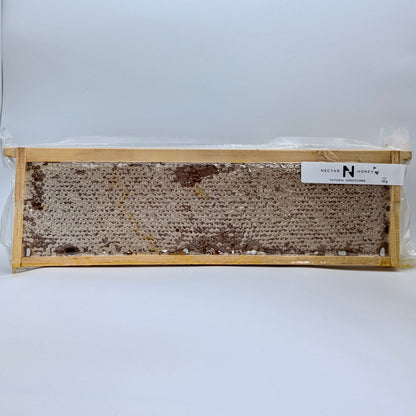 A timber frame of raw honeycomb with beeswax cells, sealed in a vacuum-sealed plastic packaging with a white label on top.